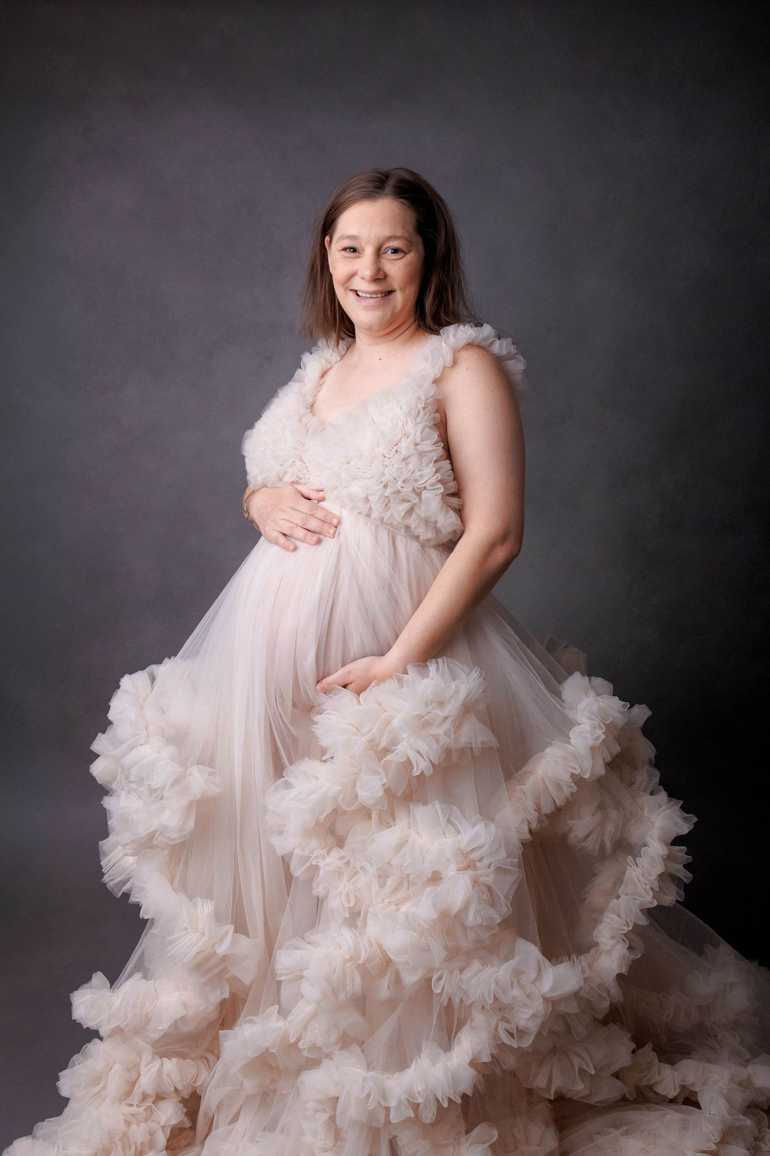Pregnant woman smiling in a flowing ruffled blush tulle maternity gown, posing in a professional studio portrait.