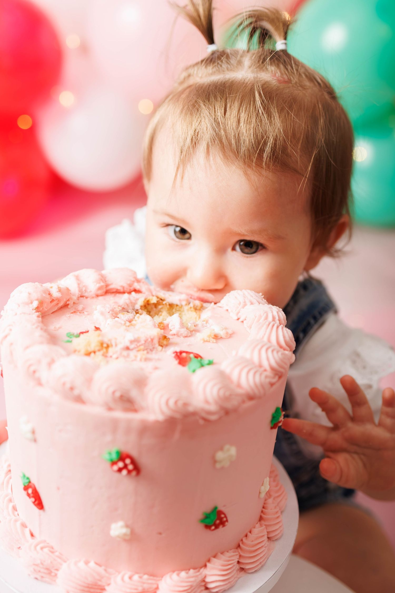 A baby girl biting into a pink birthday cake decorated with strawberries and swirl frosting at her first birthday party.
