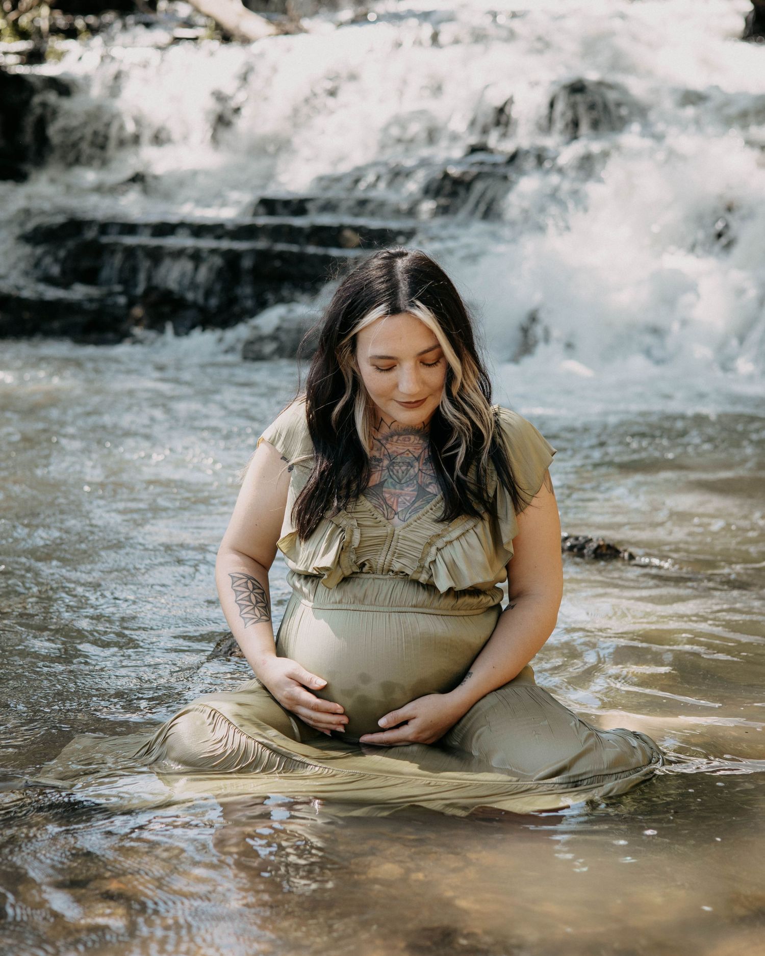 A maternity photo of someone sitting by the water wearing a sage green dress.