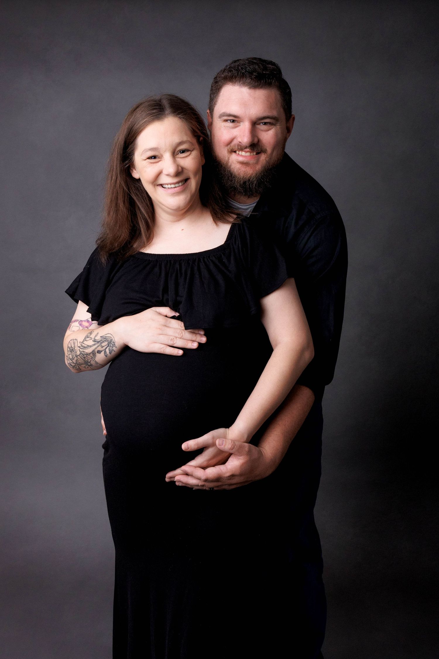 Smiling pregnant couple in black outfits pose together in a studio, man embracing woman from behind cradling her baby bump.