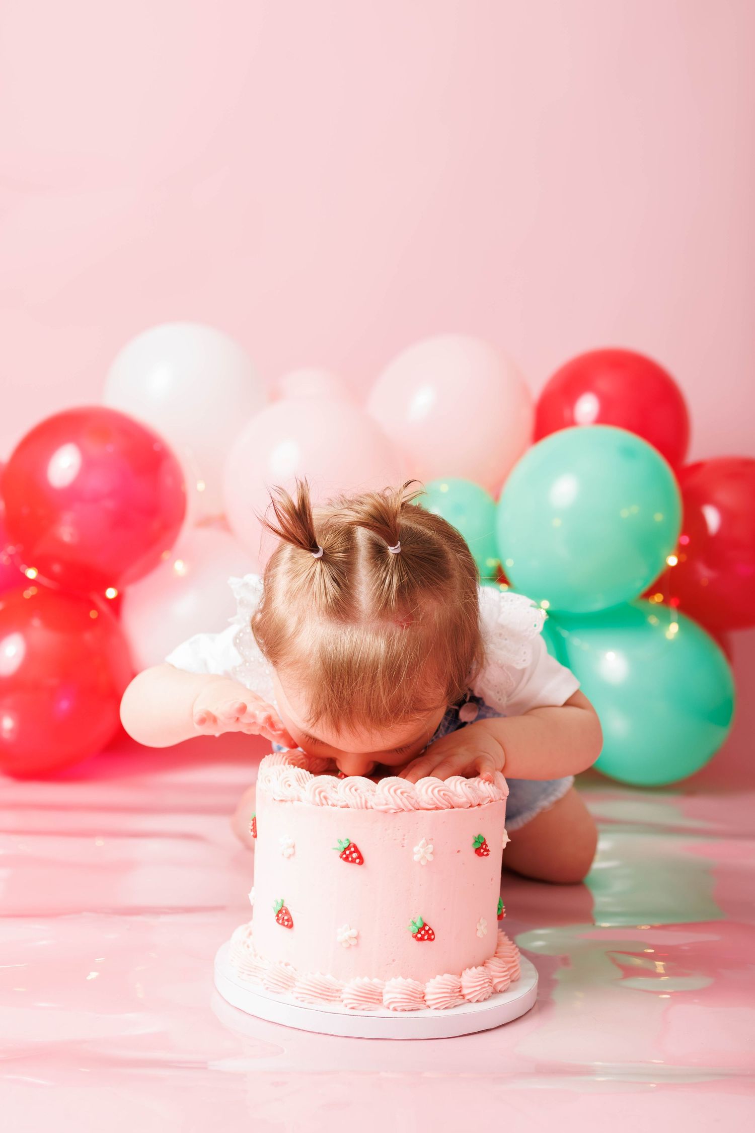 Toddler girl smashing face into pink strawberry birthday cake surrounded by colorful balloons on pink background.