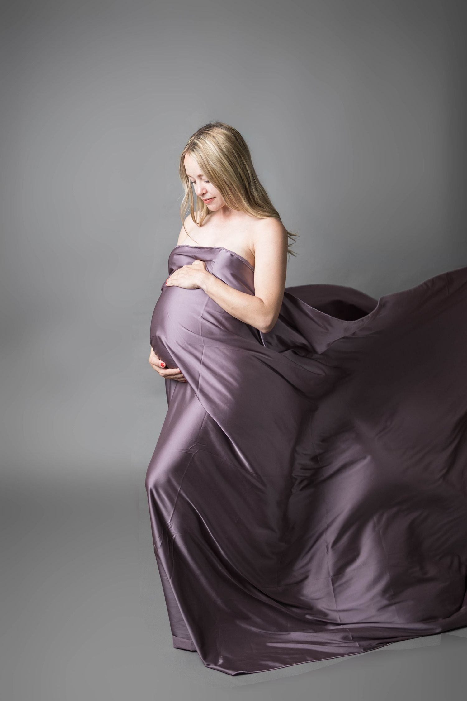 Pregnant woman in flowing purple satin fabric draped elegantly, cradling her baby bump against a gray studio background.