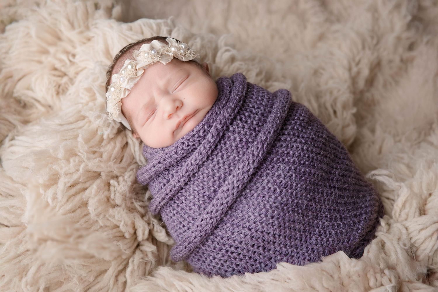 Sleeping newborn wrapped in purple knit wrap on cream textured backdrop.