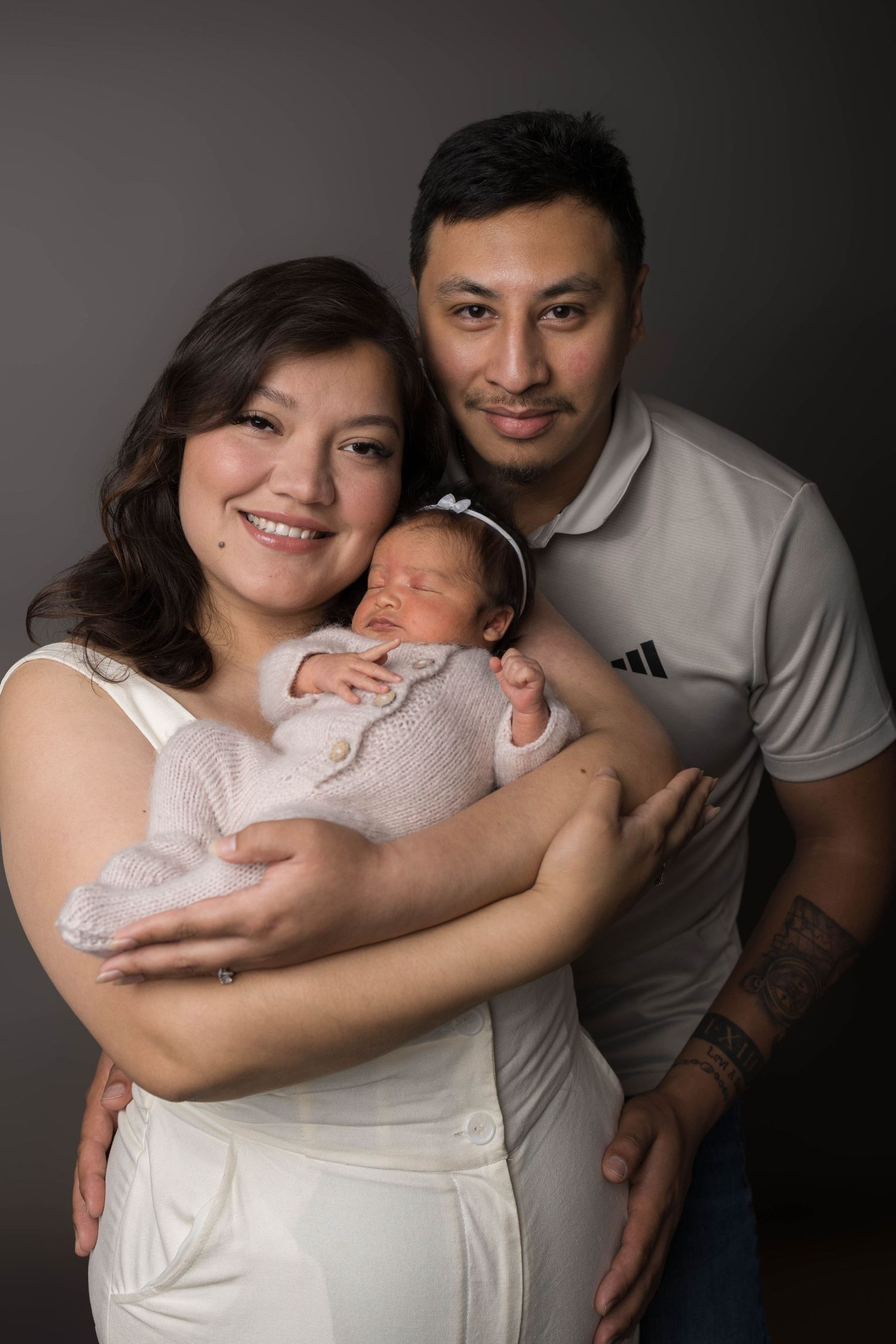 A smiling couple proudly holds their sleeping newborn baby dressed in pink for a studio portrait photo.