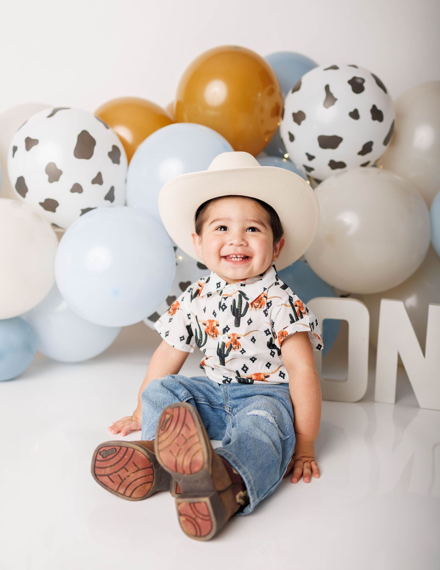 Smiling baby boy in cowboy hat, cactus shirt, and boots sits before colorful balloon backdrop at first birthday.