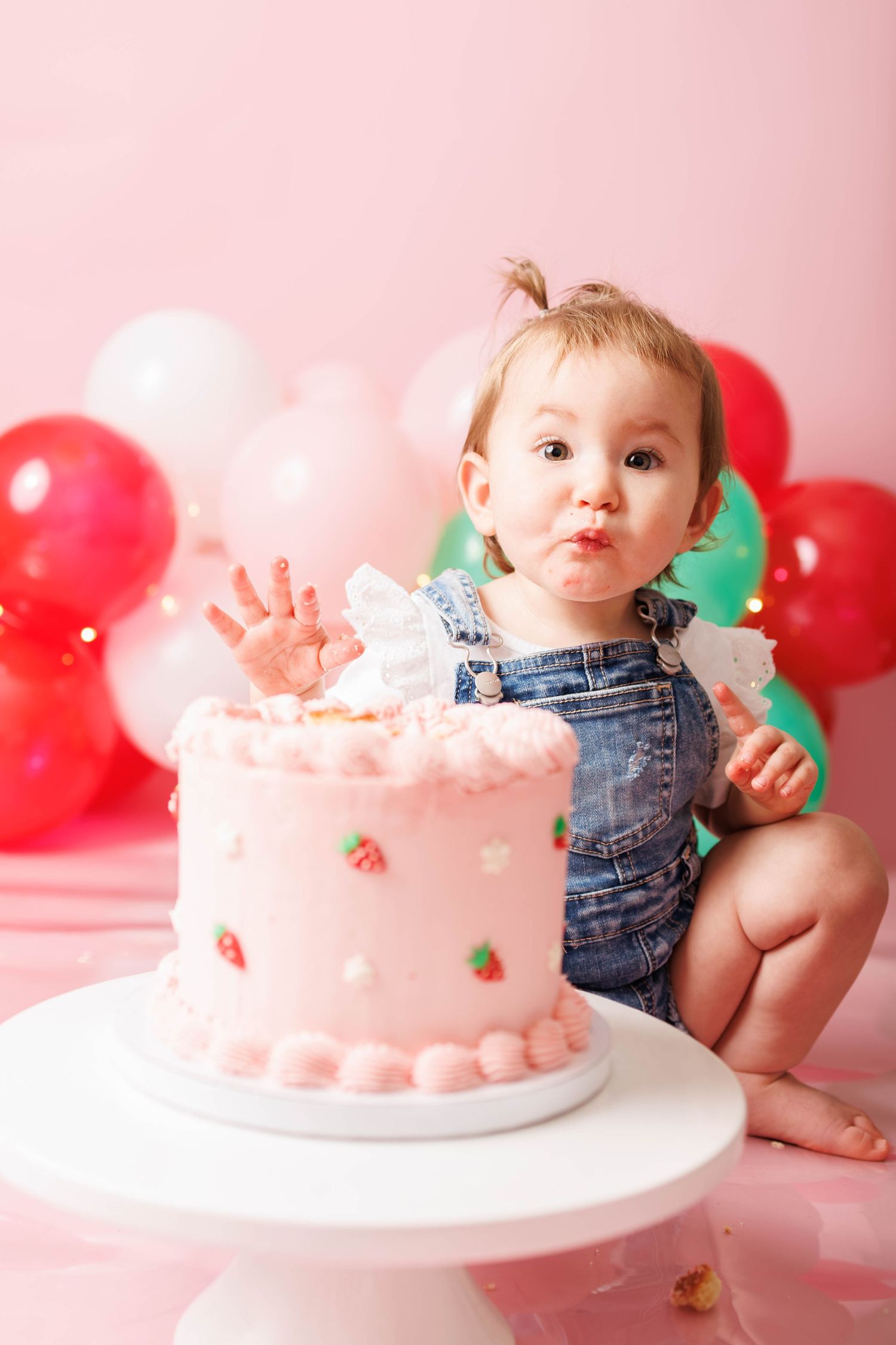 Cute baby girl in denim overalls smiling beside a pink strawberry first birthday smash cake with red balloons in background.