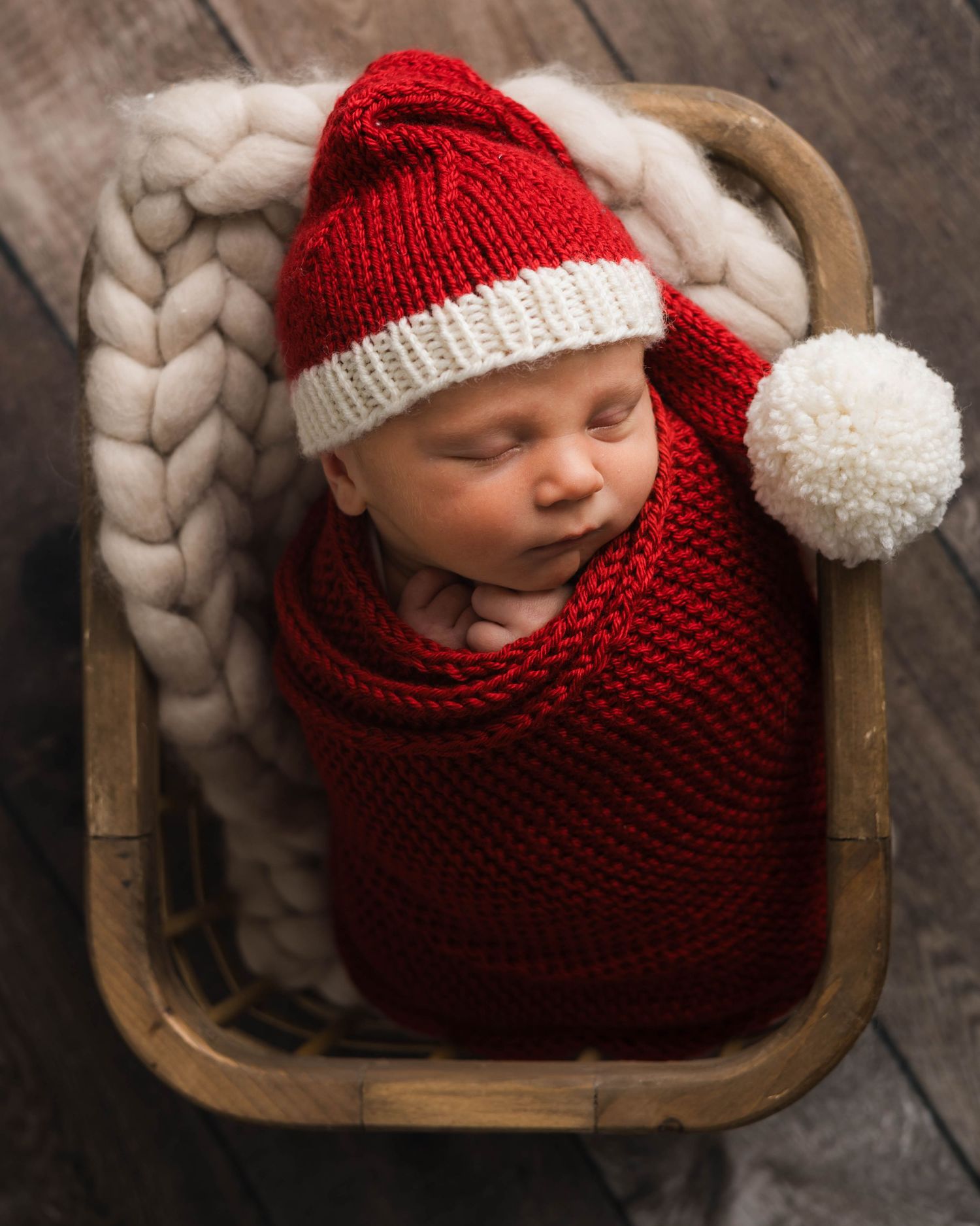 A sleeping baby wrapped in a red knitted blanket with a white pom pom hat lies in a wooden bowl against a dark background.