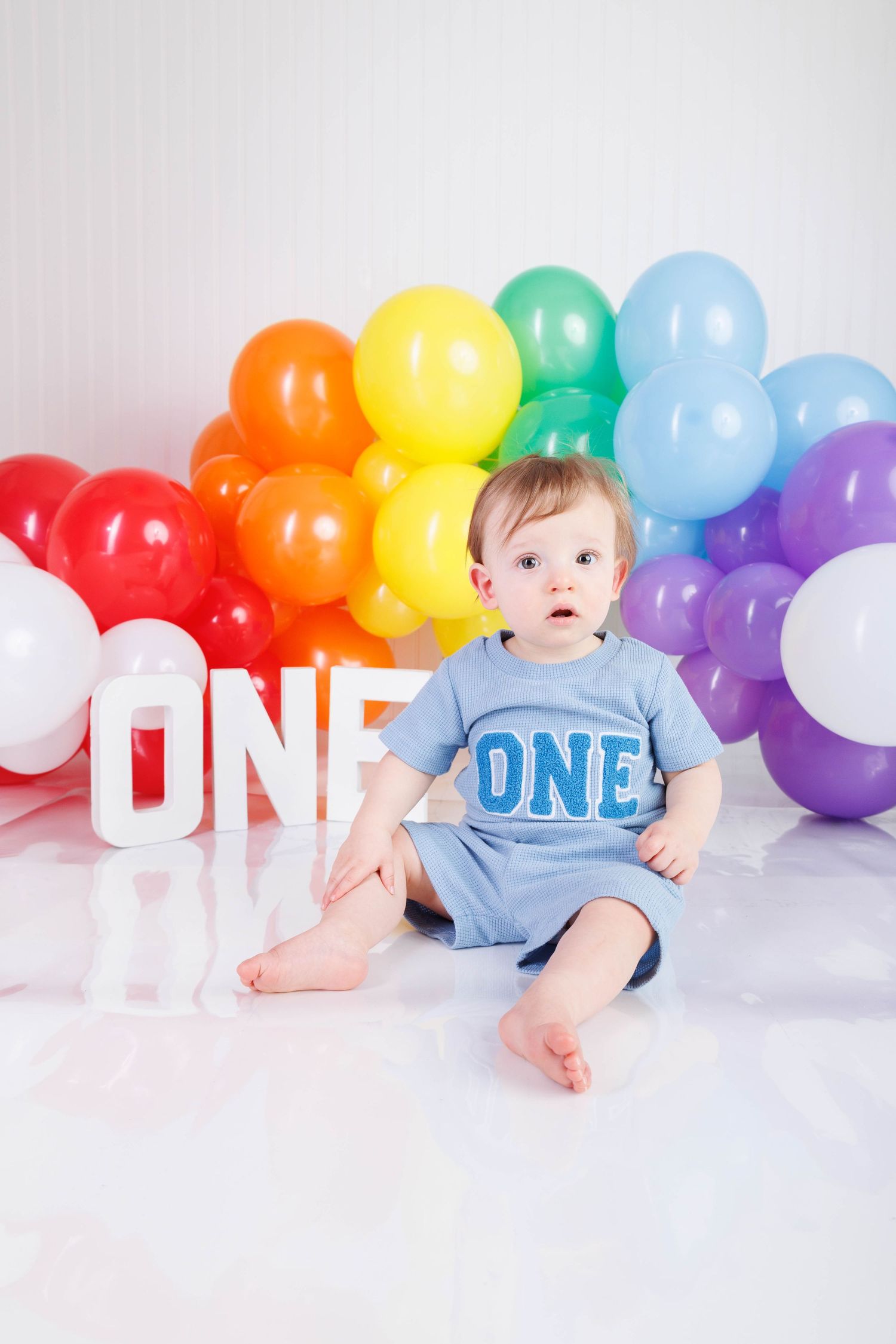 Baby in blue outfit sits among colorful rainbow balloon arch for first birthday photo shoot against white backdrop.