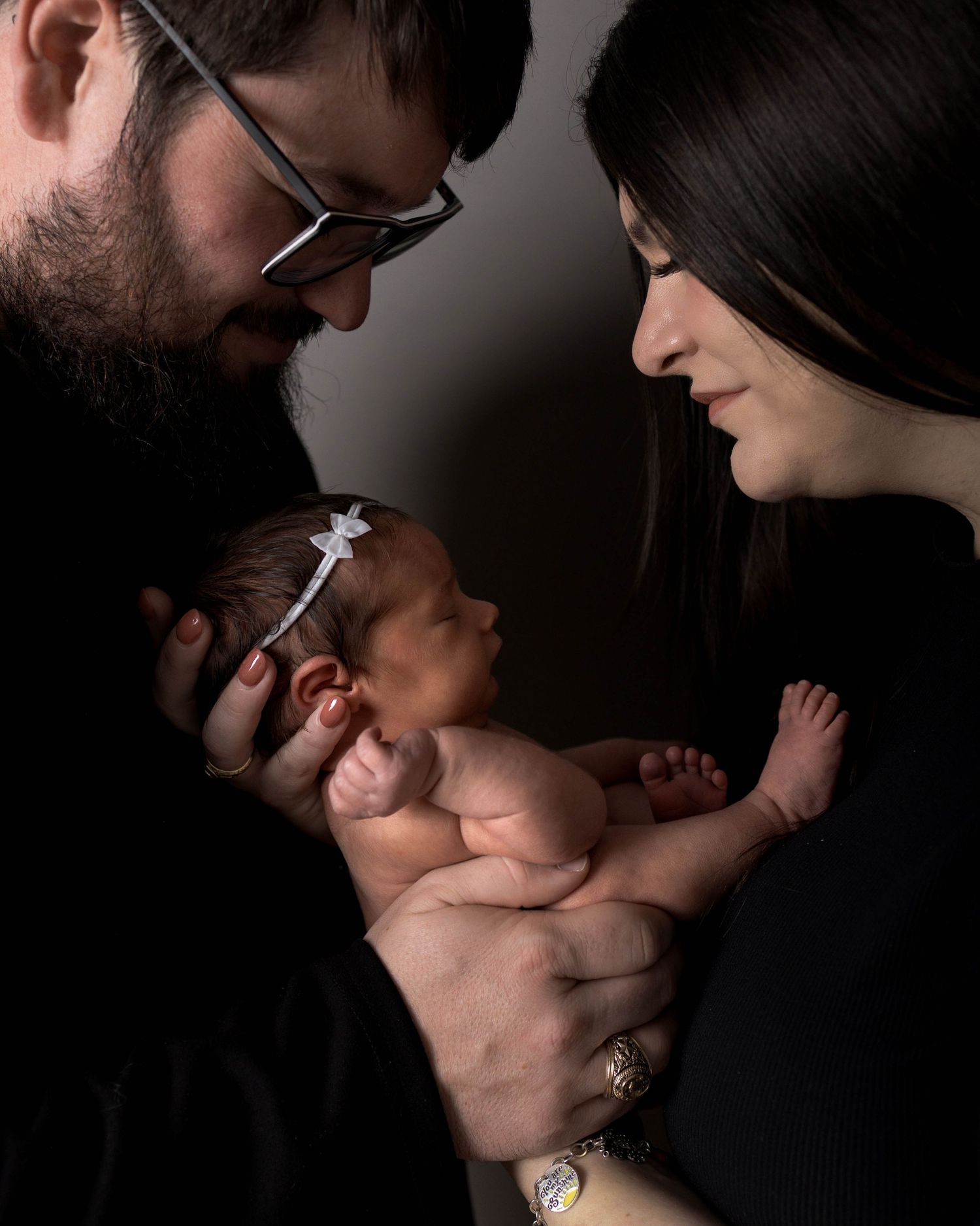 Parents in black clothing tenderly hold their sleeping newborn baby with a white bow headband in a studio portrait.