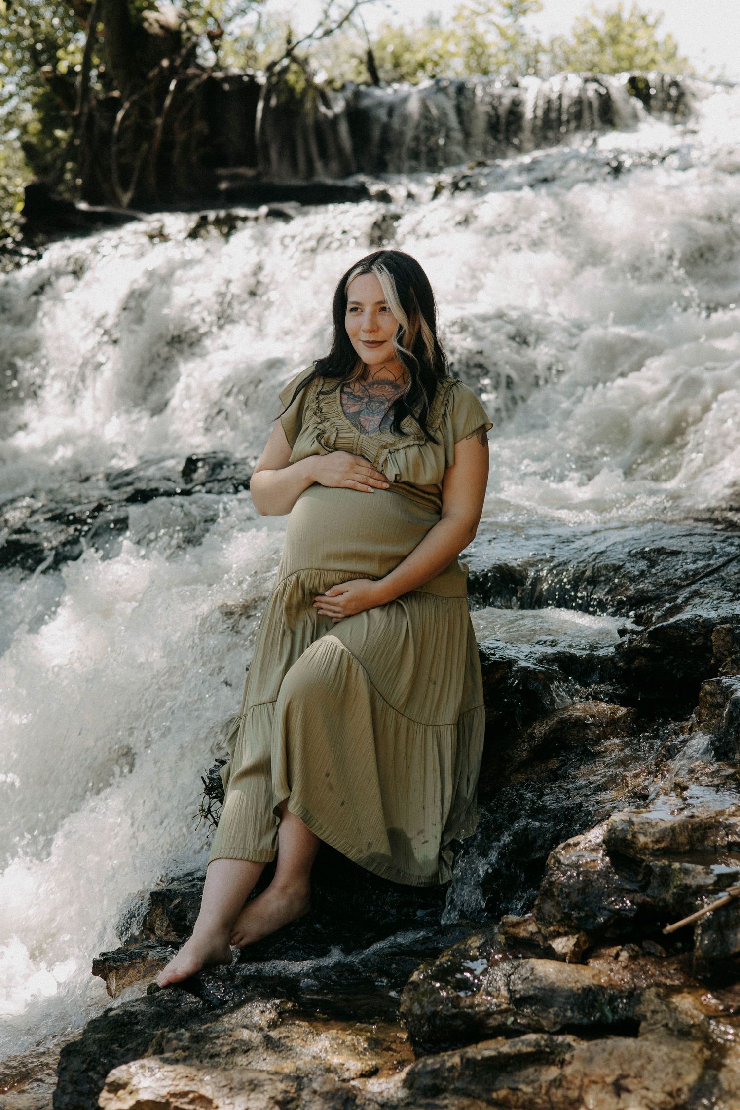 An expectant mother sits on rocks in front of a cascading waterfall wearing an olive green dress.