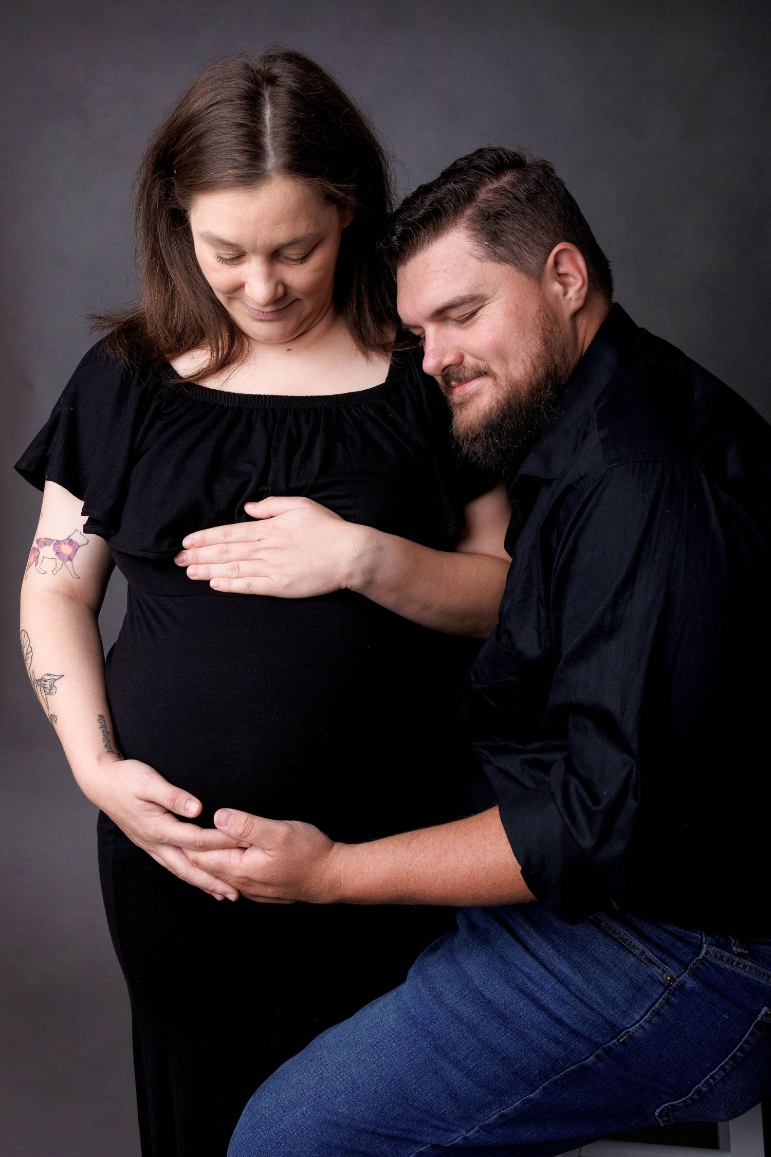 A couple in black outfits tenderly cradle a pregnant belly during a maternity photo session against a gray backdrop.