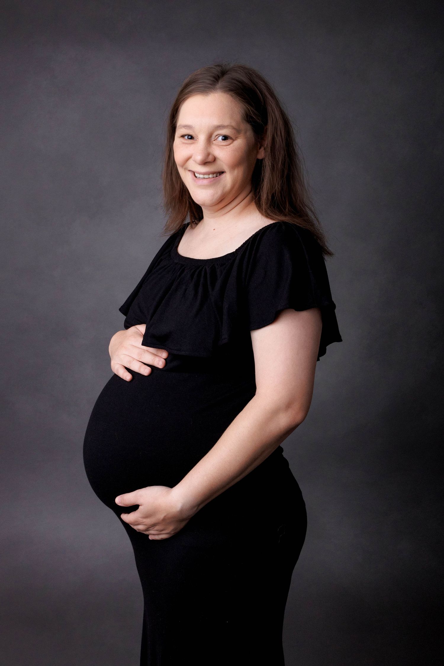 Smiling pregnant woman in black maternity dress cradling her baby bump against a gray studio background.
