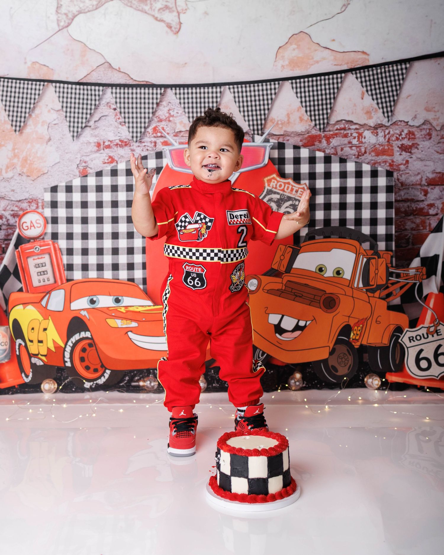 Toddler in red race car outfit stands on checkered cake at Cars-themed first birthday party with Lightning McQueen backdrop.