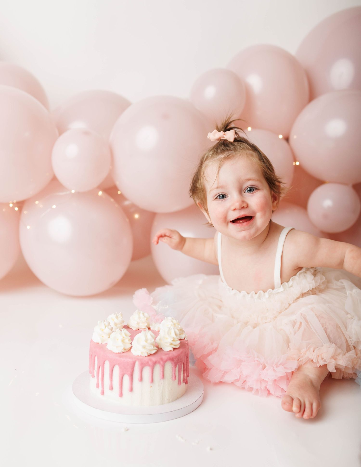 Baby girl in pink tutu smiles at first birthday cake smash with pink balloon backdrop.