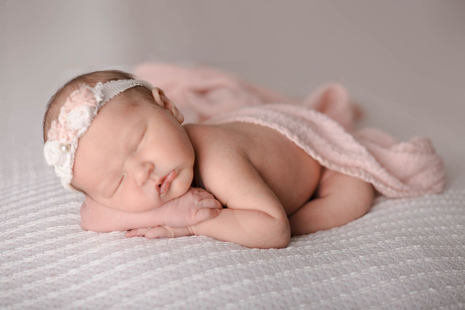 A sleeping newborn baby lies peacefully on soft white textured bedding in a side-lying position.