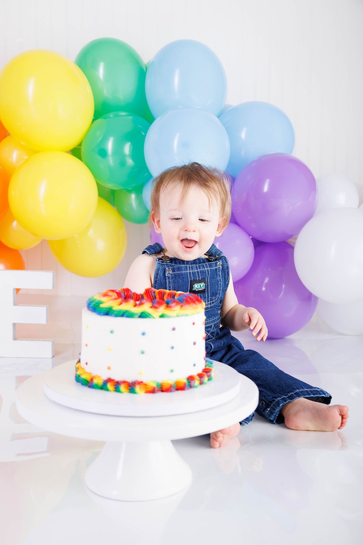 A colorful birthday celebration setup with a rainbow frosted cake on a white pedestal and festive balloons in the background.