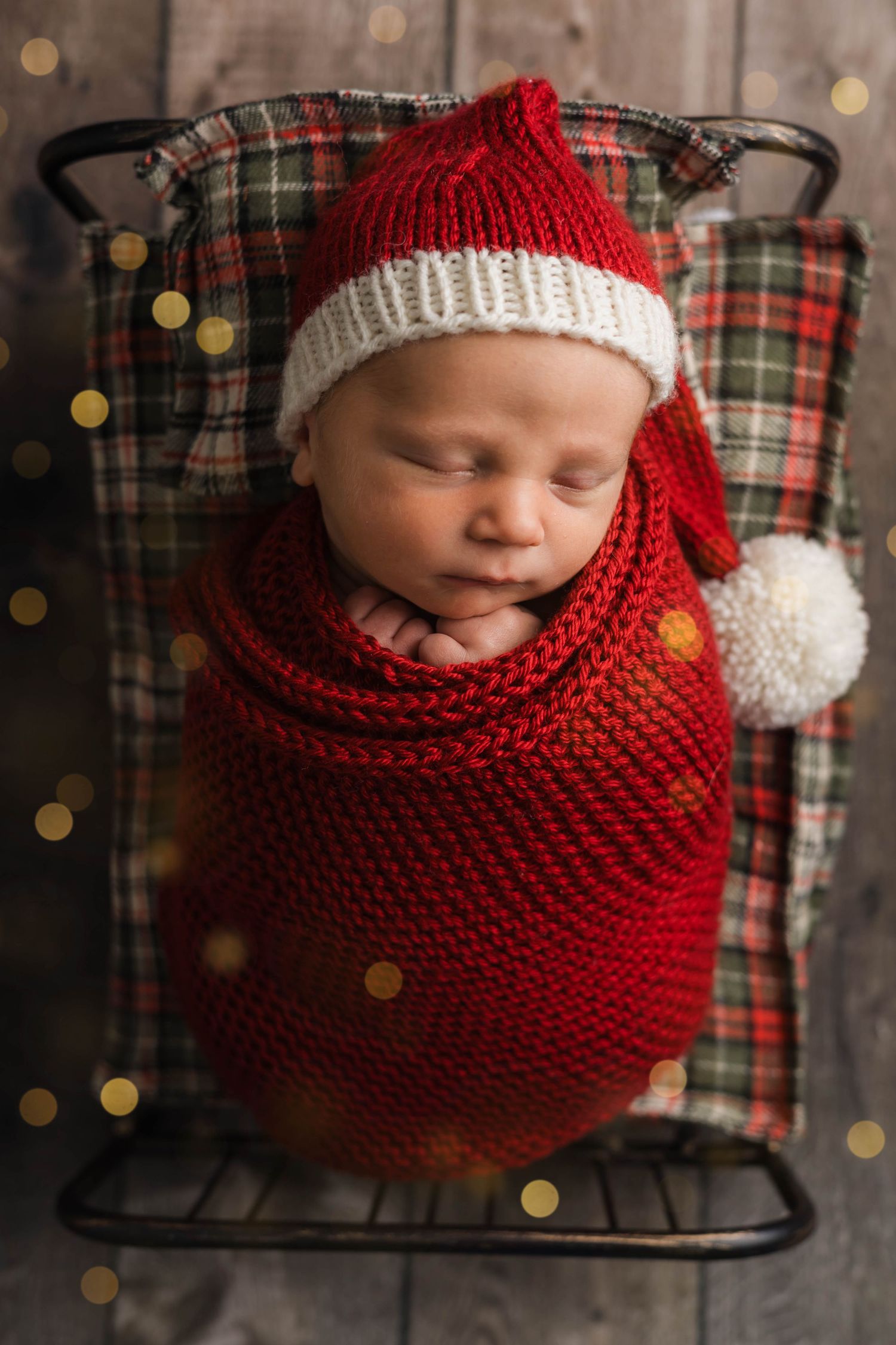 A sleeping baby wrapped in a red knitted cocoon with white pom pom details against a plaid background with bokeh lights.