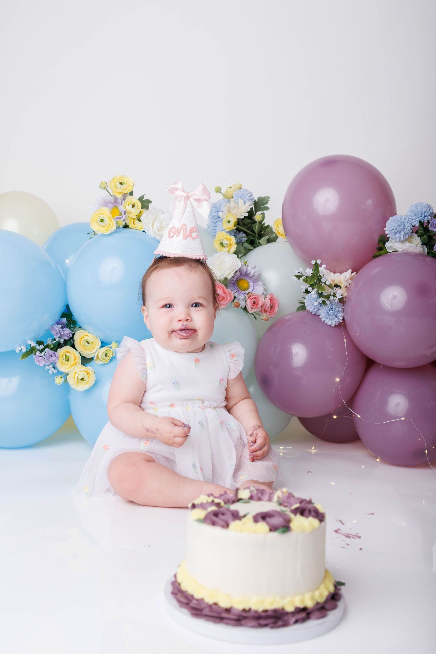 Smiling baby girl celebrating first birthday with cake, surrounded by blue and purple balloons and flowers.