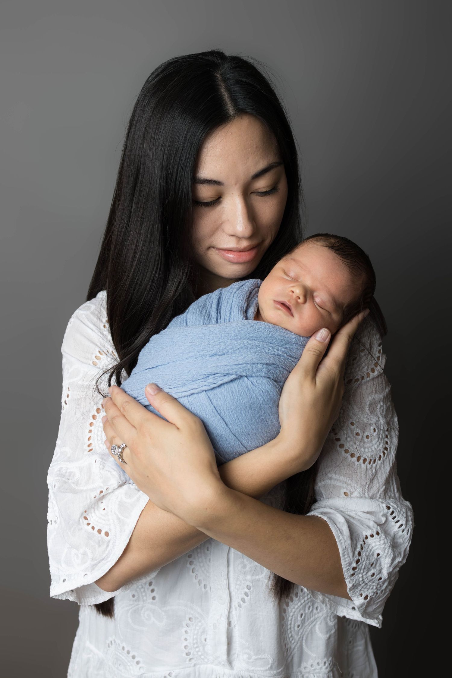 A mother in white dress tenderly holds her sleeping newborn wrapped in blue against a gray background.