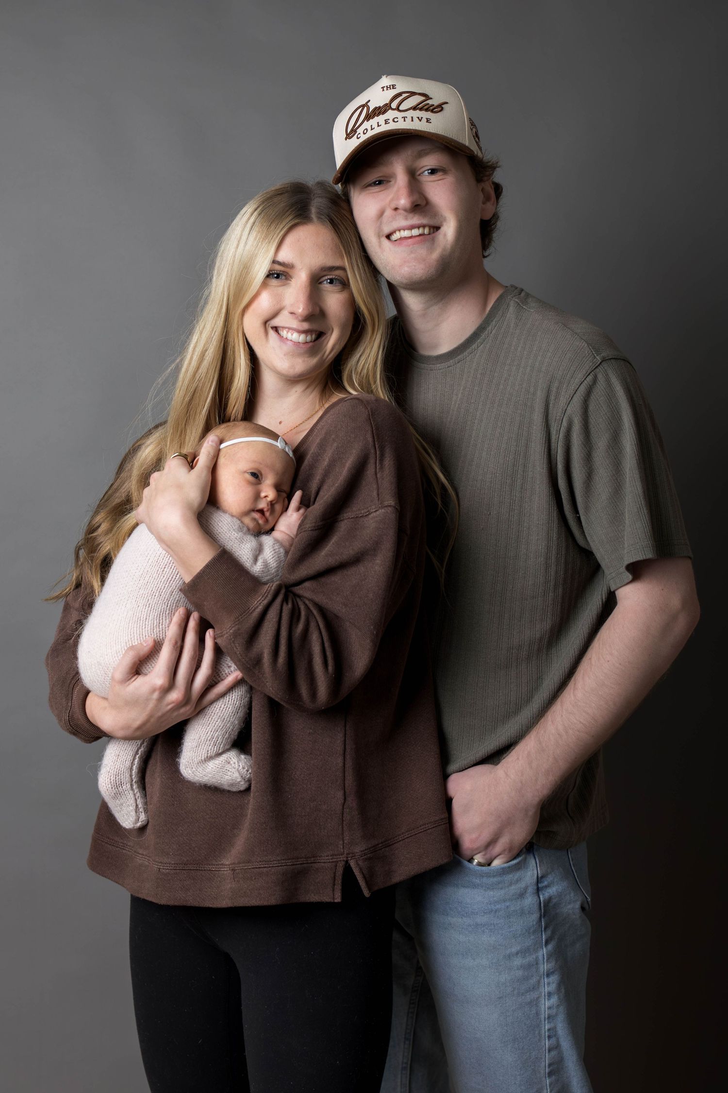 Smiling couple posing with their newborn baby in a studio portrait, woman in brown sweatshirt, man in green shirt and cap.