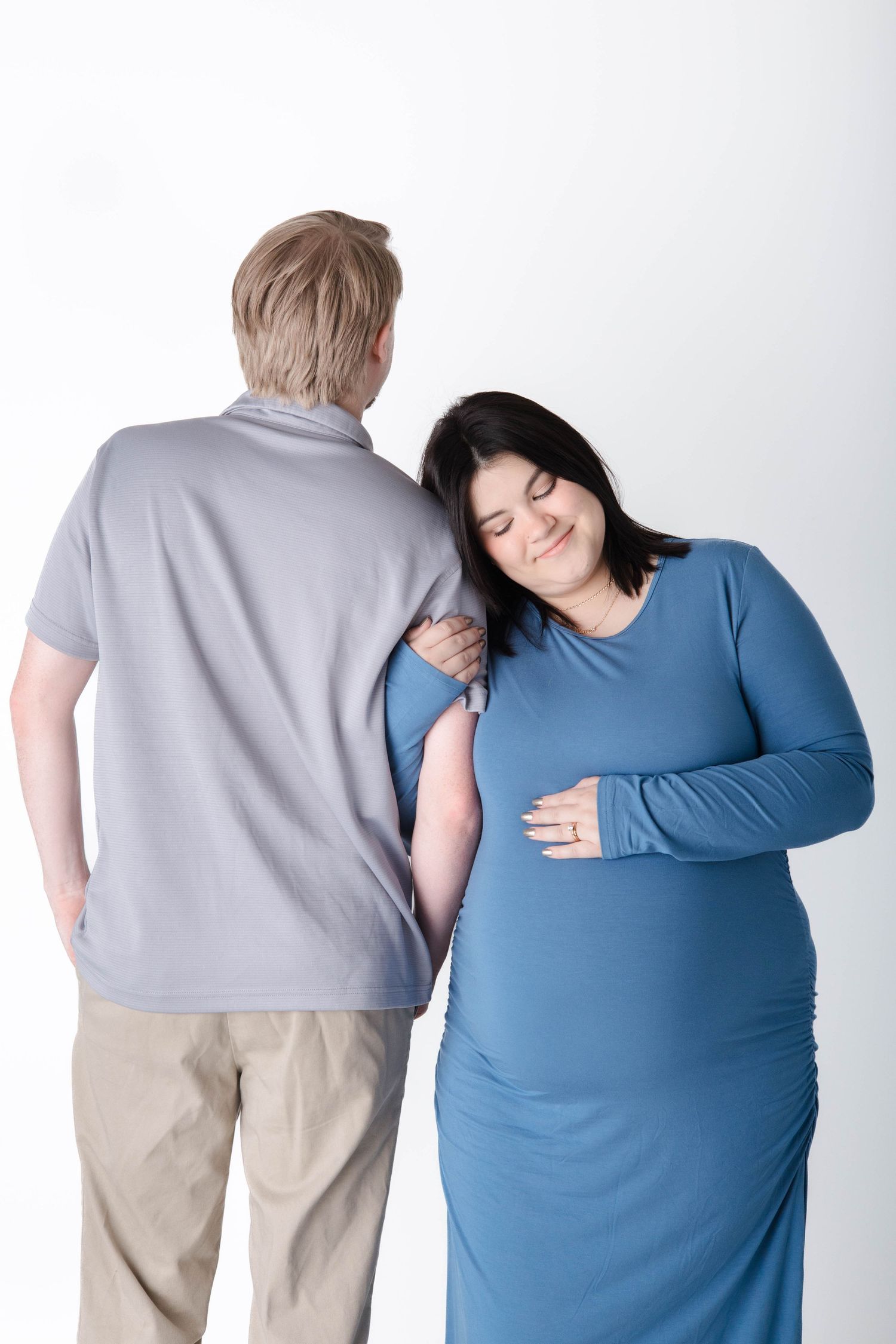 A couple shares an intimate moment as the pregnant woman in a blue dress leans against her partner in a grey shirt.
