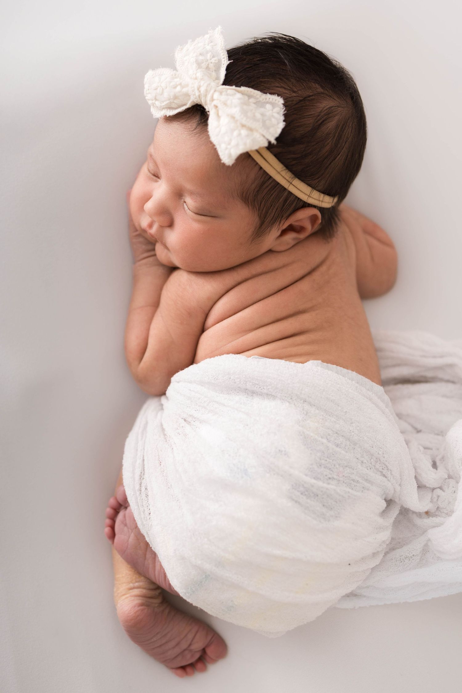 Sleeping newborn baby girl wrapped in white swaddle with bow headband on white background.