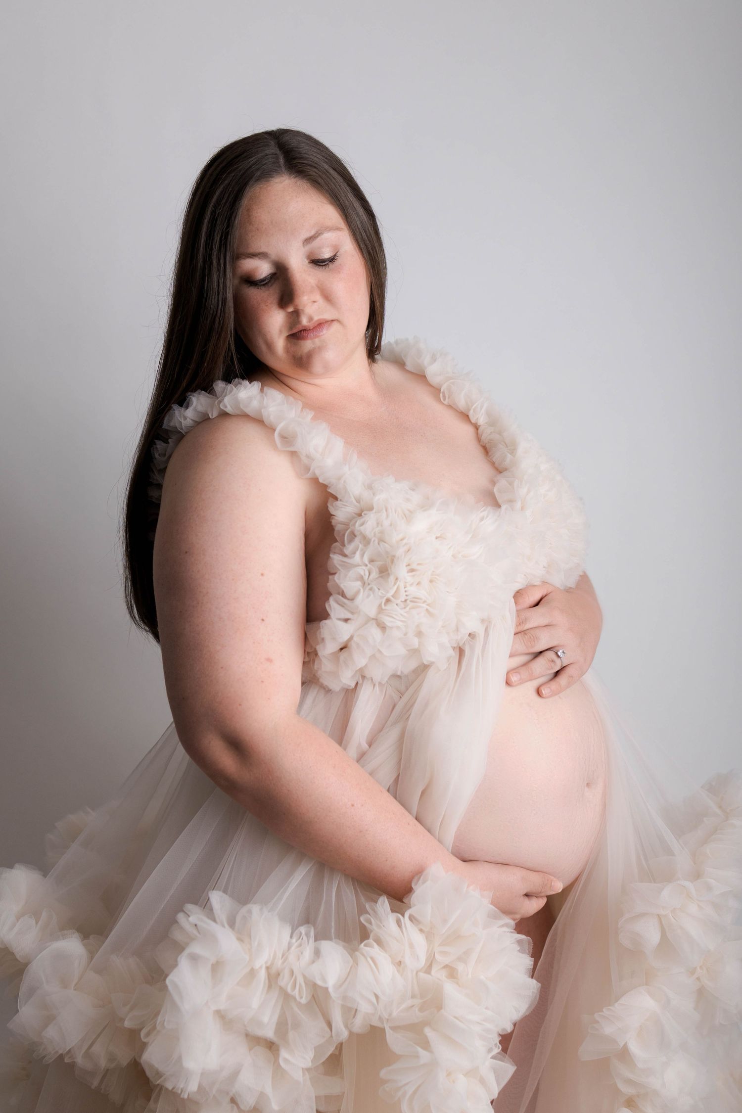 Pregnant woman in a ruffled cream tulle maternity gown cradling her bare baby bump against a white background.