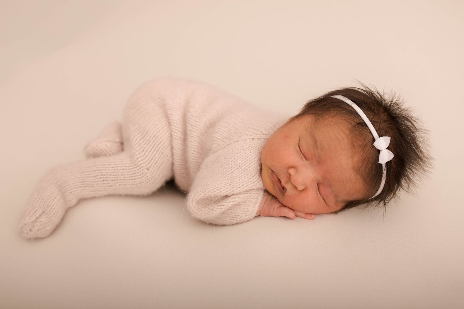 Sleeping newborn baby girl in pink knit outfit with white bow headband on soft white background.