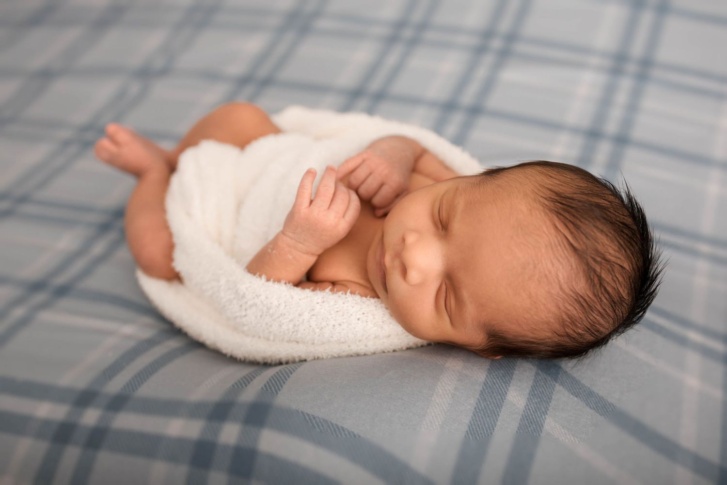 Peaceful newborn baby sleeping wrapped in white blanket on soft blue plaid background.