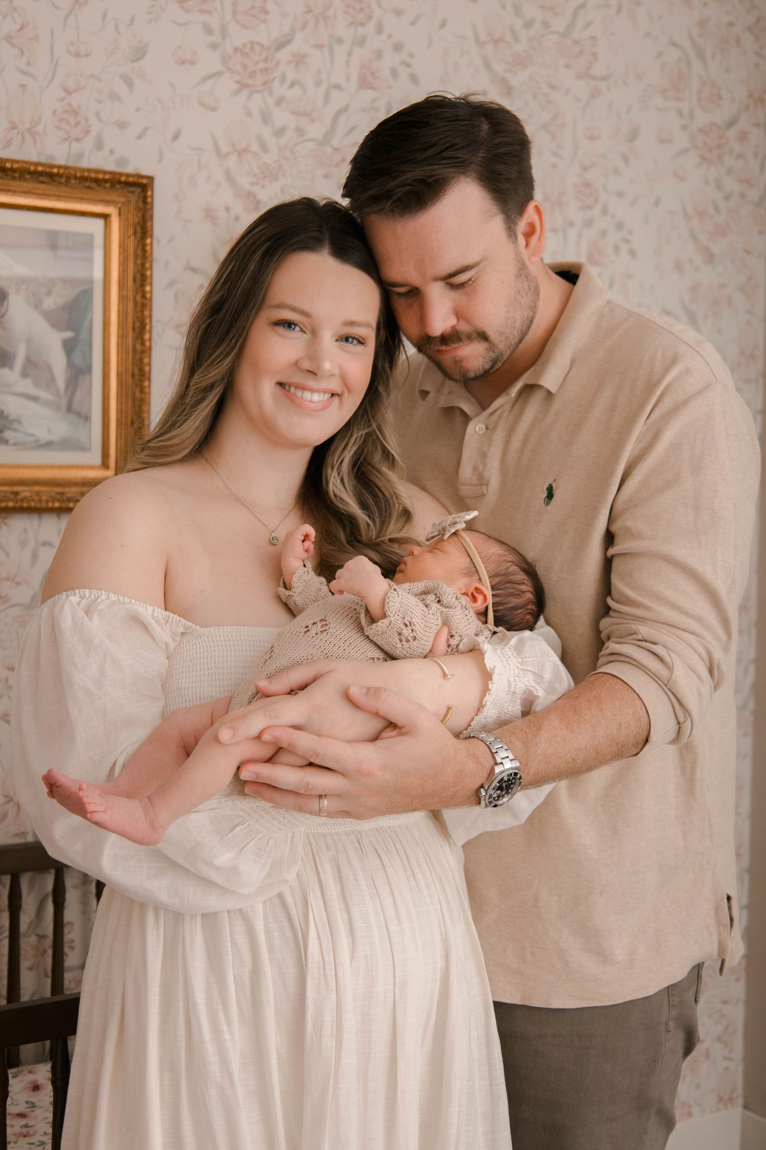 A tender family moment captured in front of floral wallpaper as parents cradle their newborn.