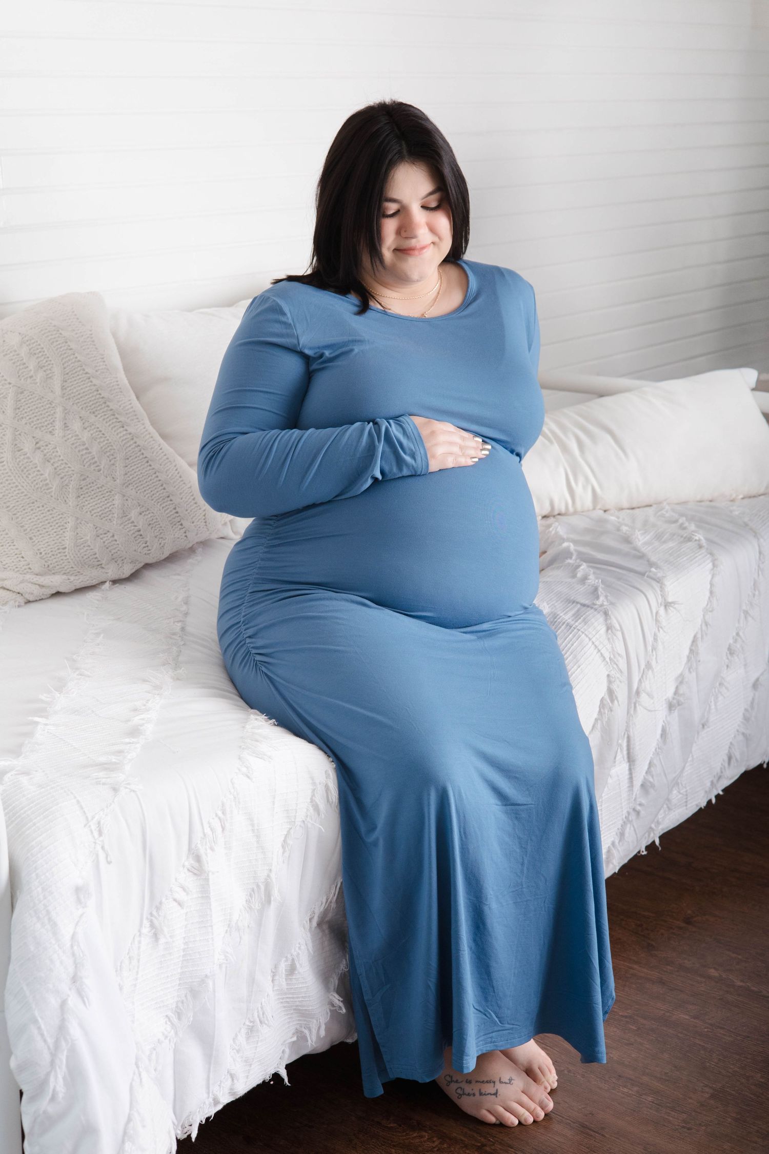 A pregnant woman in a blue dress sits on a white couch in a series of maternity photos.