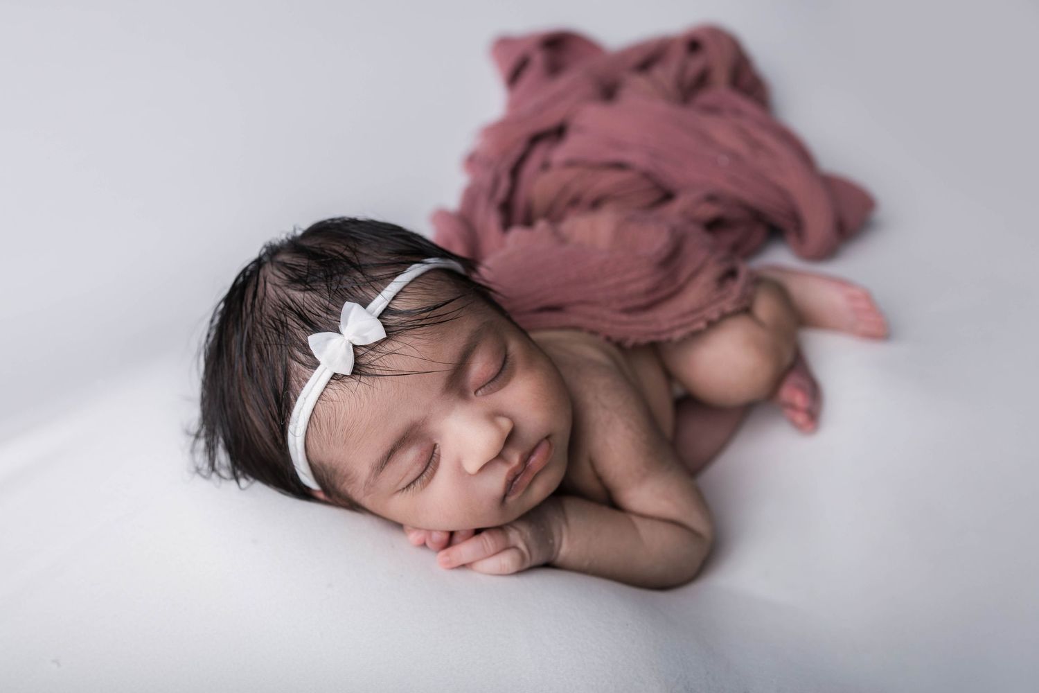 Peaceful newborn baby girl sleeping on white surface wearing a white bow headband with a pink wrap.