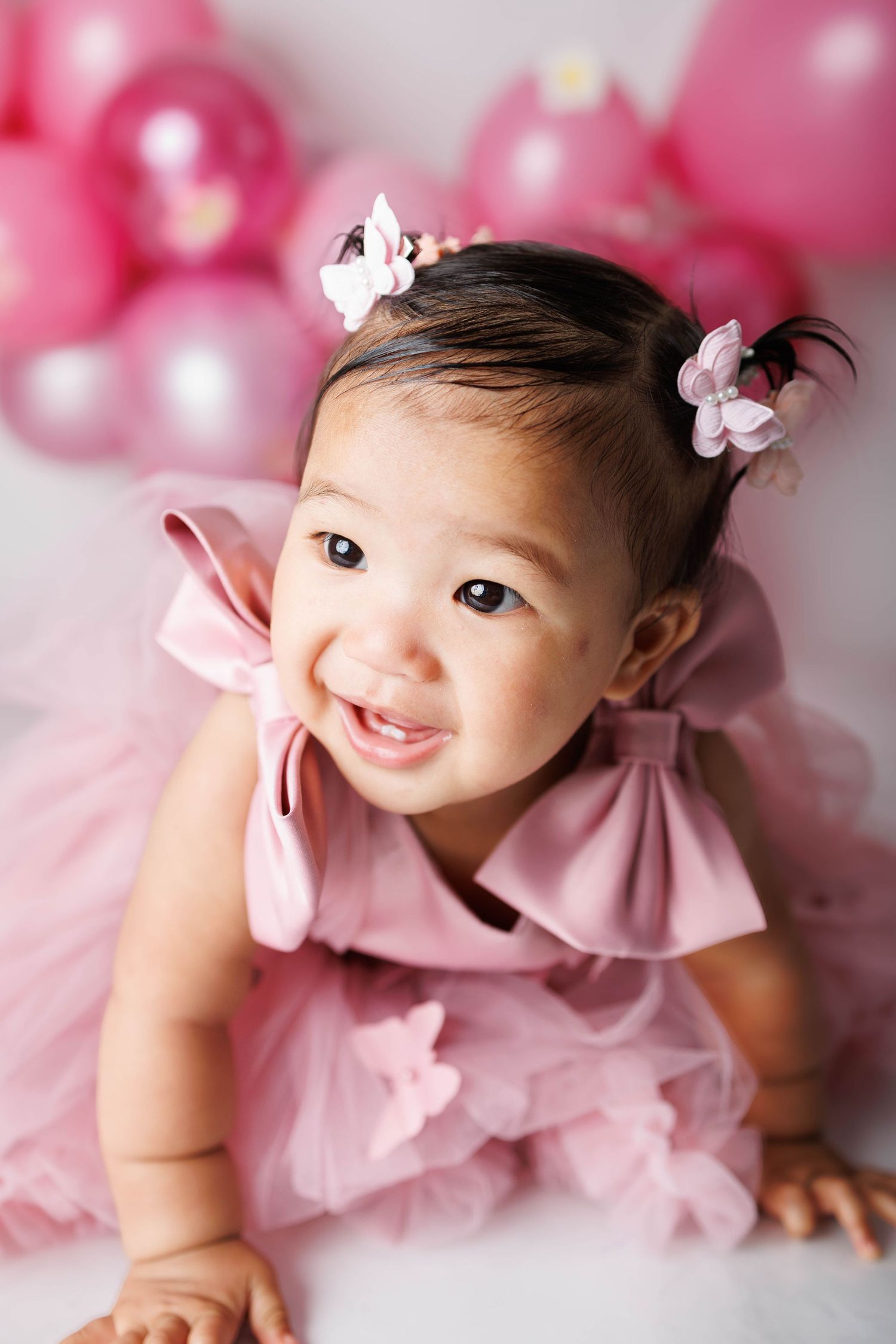 Smiling baby girl in pink tutu dress with hair clips, surrounded by pink balloons in a first birthday photo shoot.