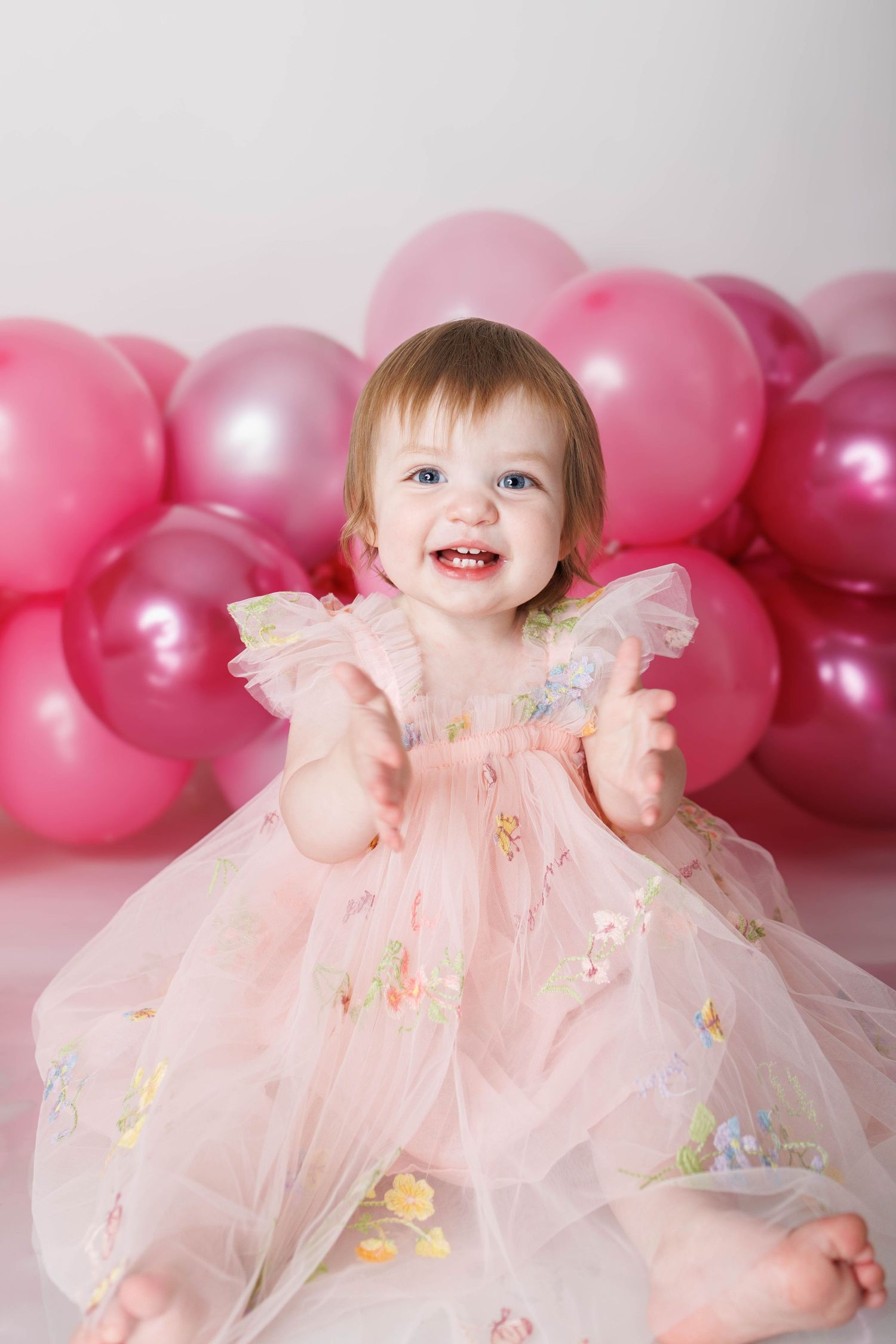 Smiling baby girl in pink tulle dress sits among pink balloons during first birthday cake smash photoshoot.