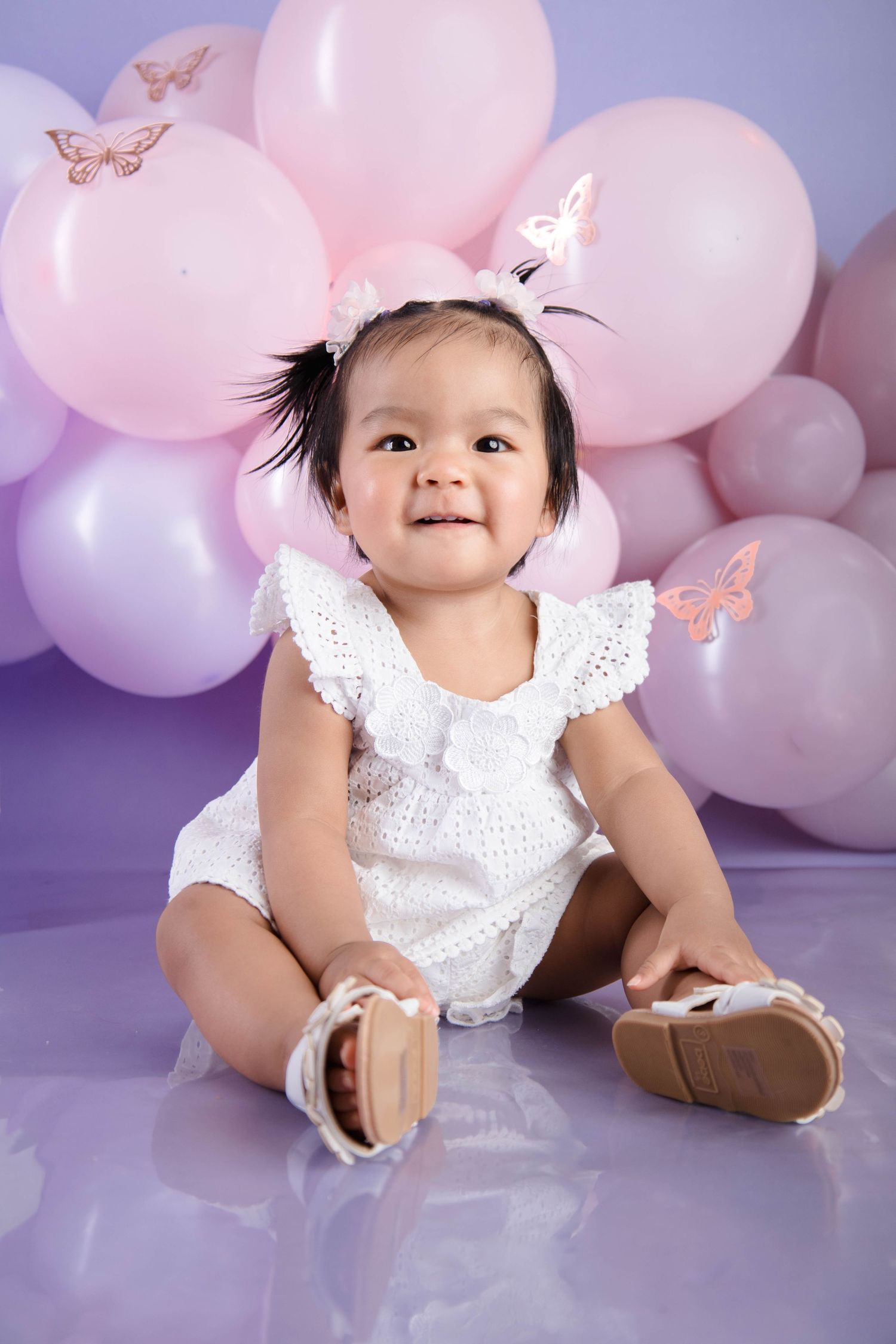 Baby in white eyelet romper sitting against purple and pink balloon backdrop with butterfly decorations.