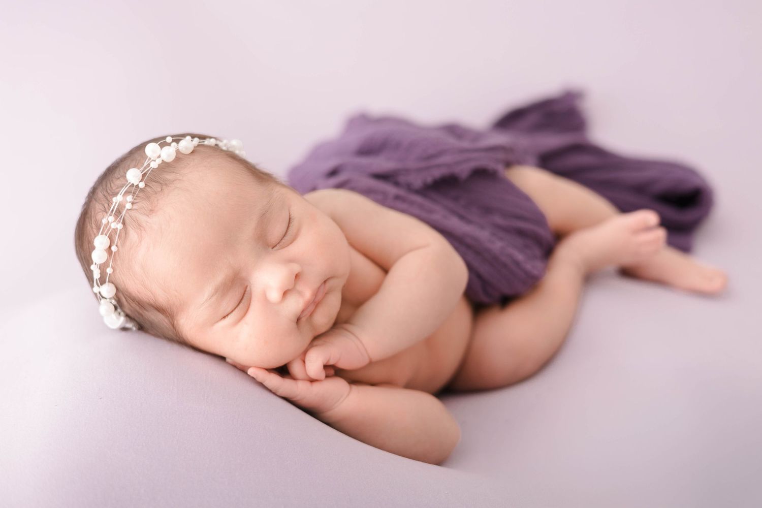 A sleeping newborn baby wearing purple fabric lies peacefully on a soft white blanket during a professional photoshoot.