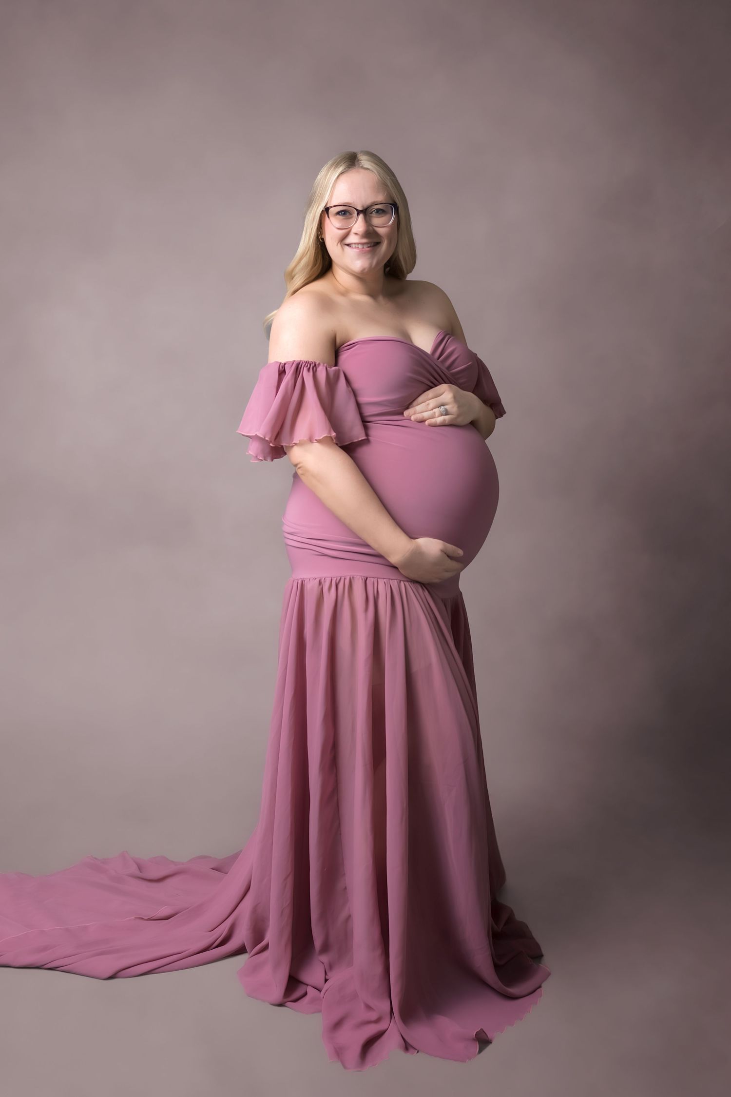 Smiling pregnant woman in elegant off-shoulder dusty rose maternity gown with train, wearing glasses, cradling baby bump.