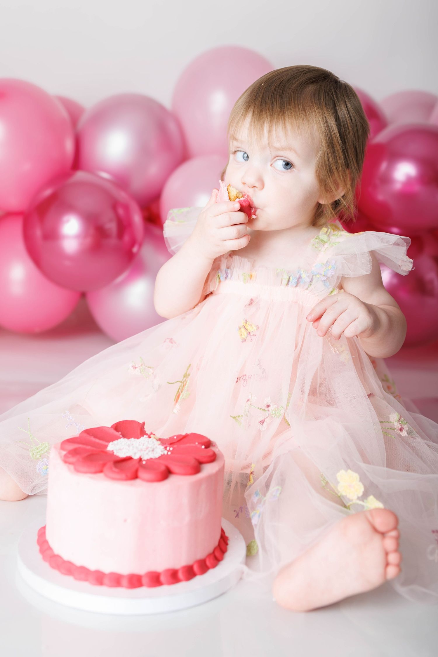 Toddler girl in pink tulle dress eating cake at first birthday party with pink balloons in background.