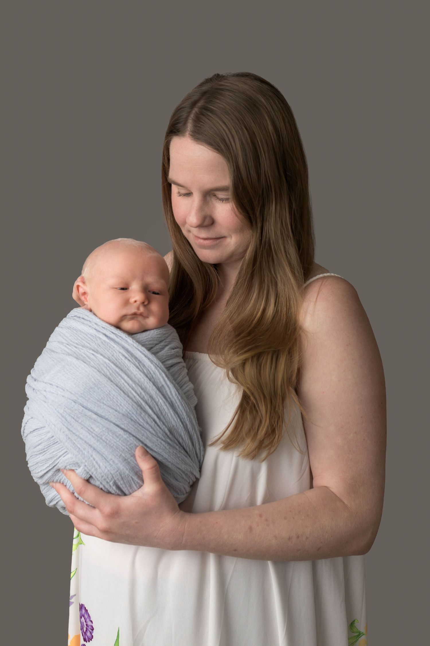A mother tenderly holds her swaddled newborn baby wrapped in a soft blue blanket against a gray studio background.