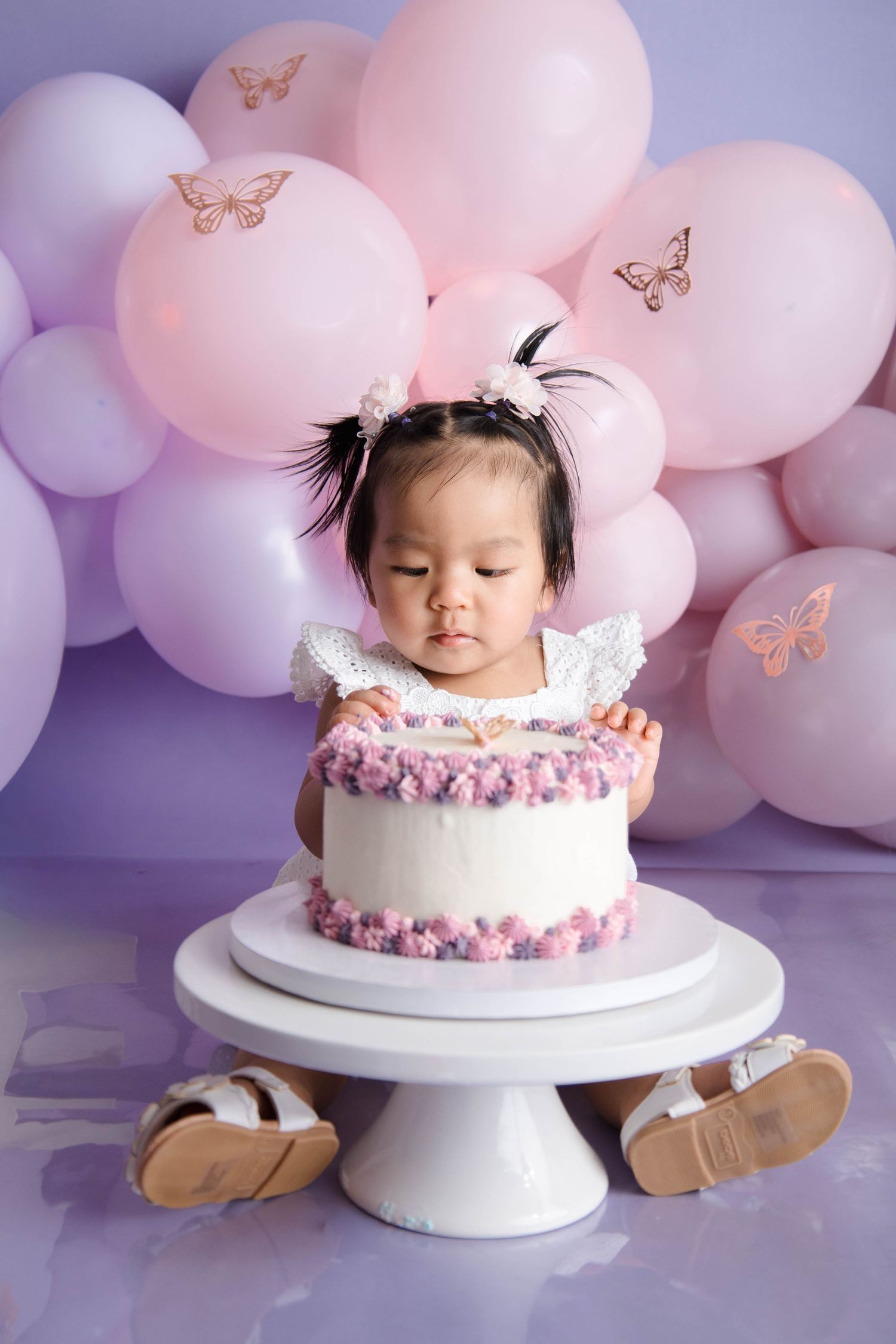 Pink and purple balloon backdrop with white cake stand for first birthday celebration with butterflies and pastel decorations.