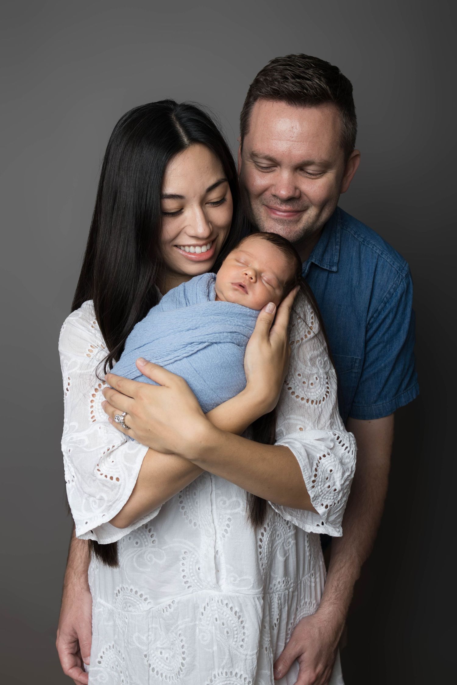 Happy parents smiling and holding their sleeping newborn baby wrapped in a blue swaddle during a studio portrait session.