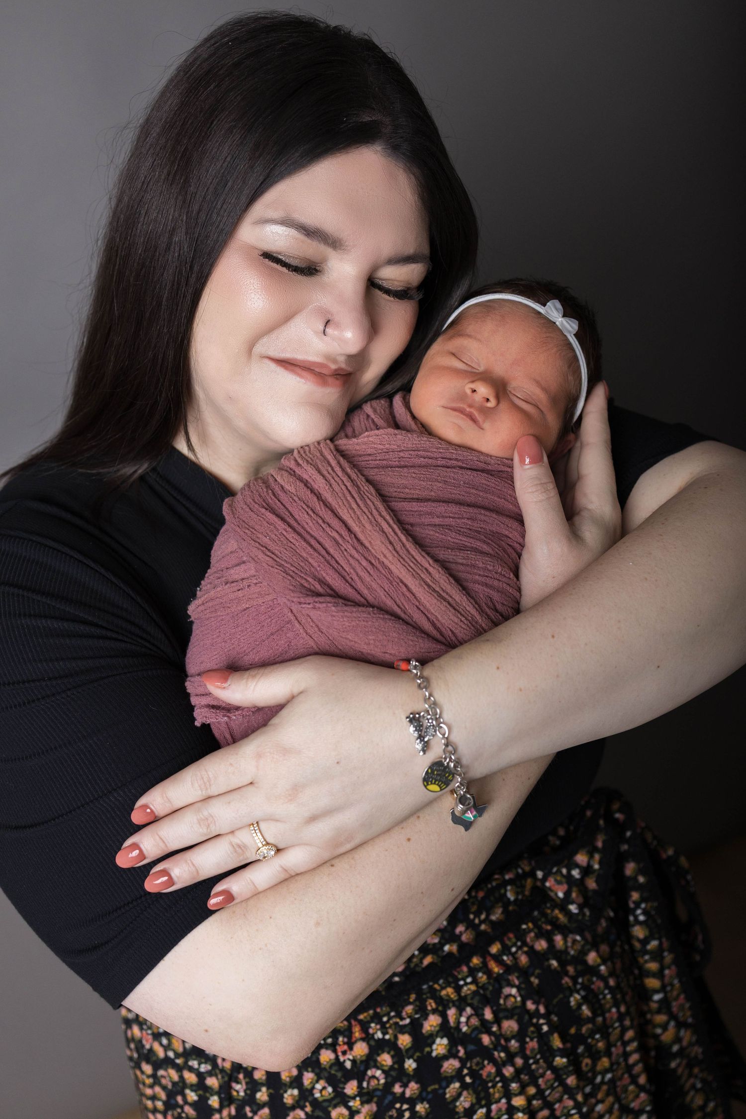 A young mother with dark hair tenderly holds her swaddled newborn baby wrapped in a mauve cloth with a bow headband.