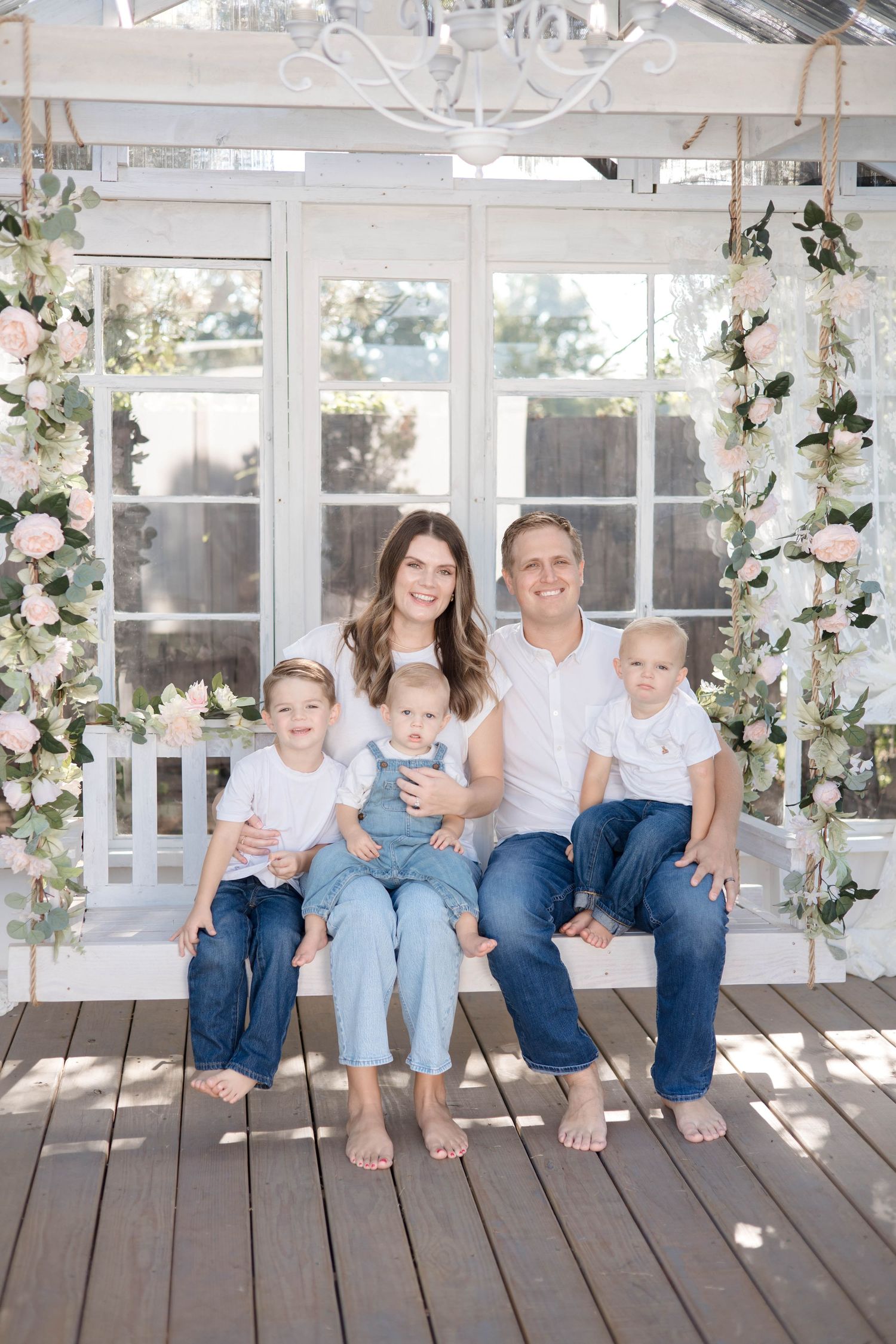 A group sits together wearing coordinated denim and white outfits on a sunlit porch decorated with floral garlands.