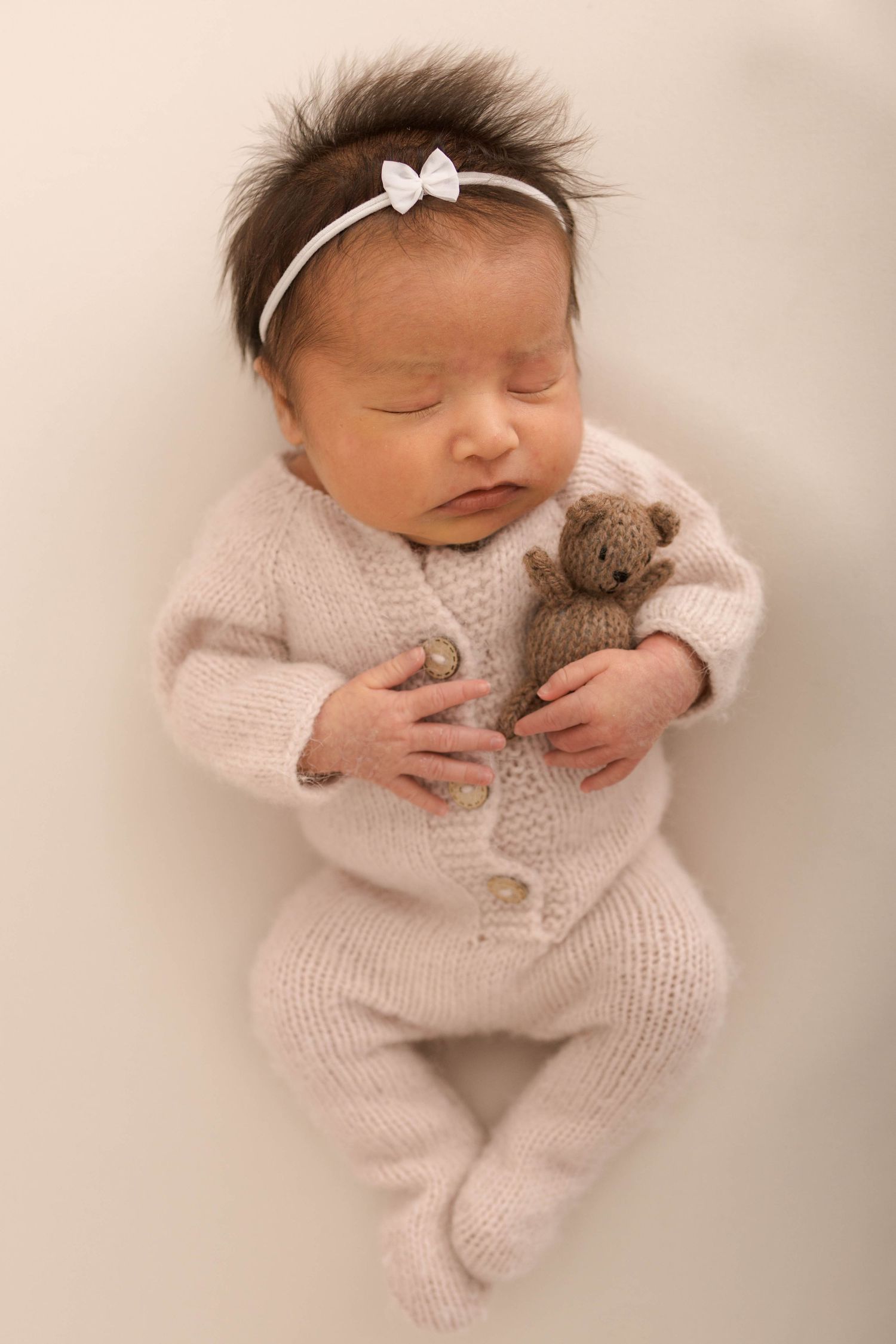 Sleeping newborn baby girl in knit outfit holding tiny teddy bear, wearing white bow headband.