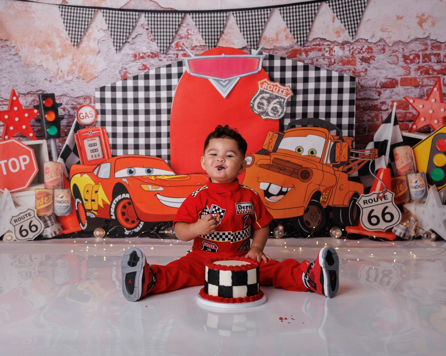 Baby boy in red outfit sits beside a checkered cake at a Cars-themed first birthday cake smash photo shoot.