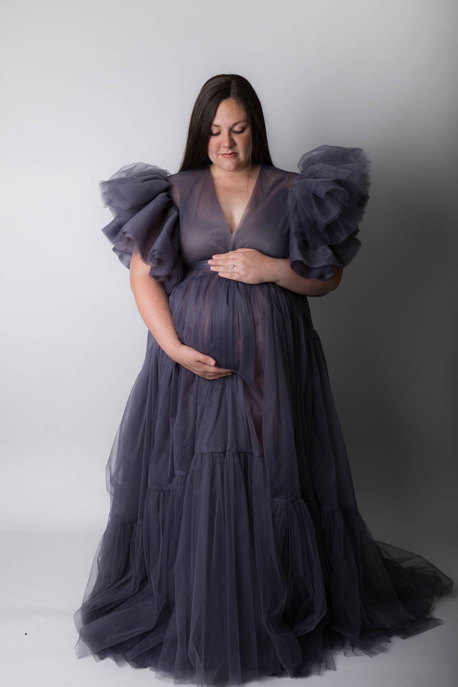 Pregnant woman in elegant gray tulle maternity gown with ruffled sleeves, cradling baby bump in studio.