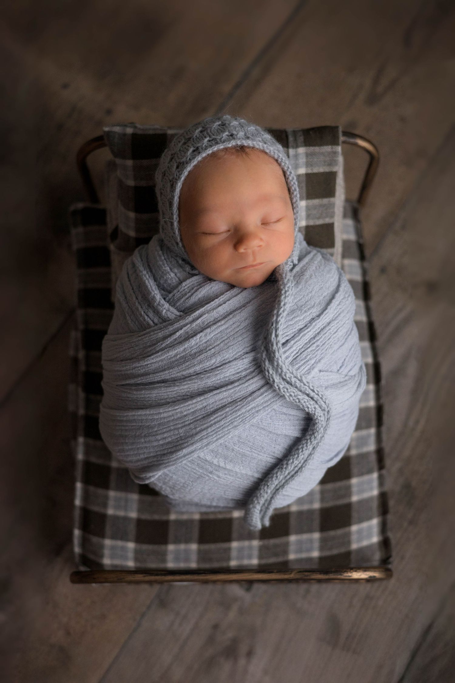 A sleeping newborn baby swaddled in soft gray fabric lying on a plaid blanket shown from different angles.