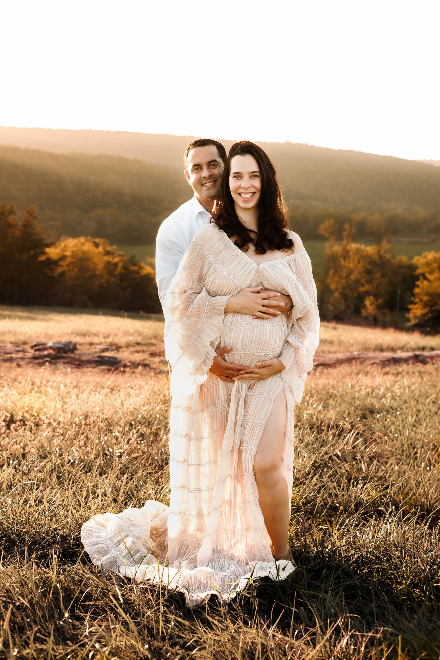 Romantic sunset photoshoot in a golden field with couple wearing white flowing dresses against mountain backdrop.