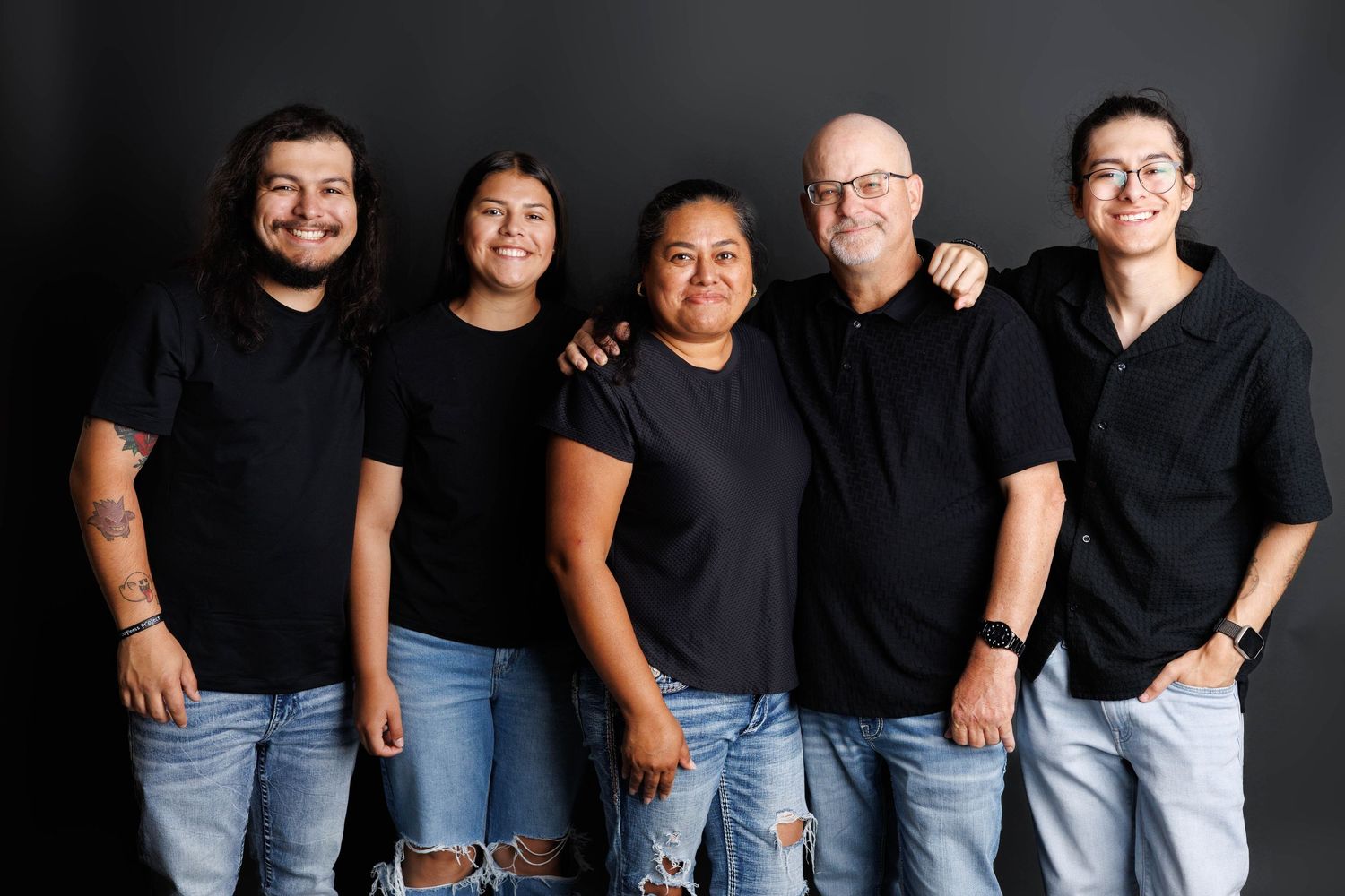 Five people wearing coordinated black t-shirts and jeans pose together against a dark background.