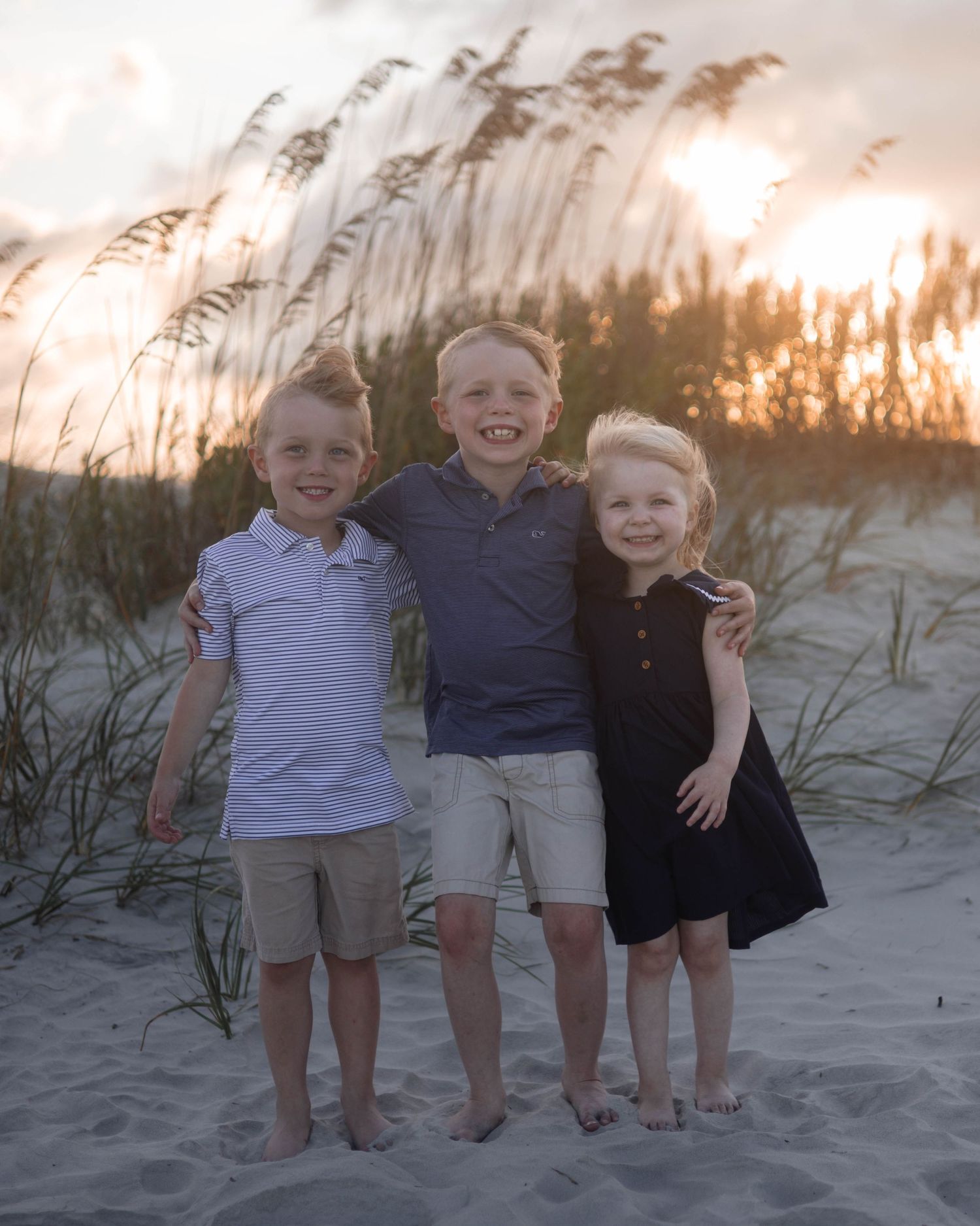 Three young children stand together barefoot on sand dunes at sunset with sea grass in the background.